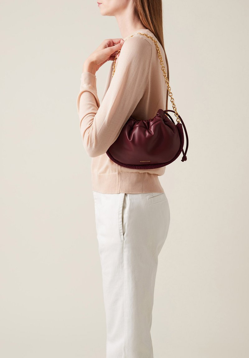 Burgunderfarbene Ledertasche mit gerafftem Oberteil, goldener Kettenriemen und gewebten Akzenten. Kombiniert mit einem hellen Pullover und hellen Hosen.