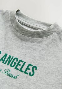 Pelēks krekls ar ribota apakšmalu un zaļu tekstu, kas lasa "LOS ANGELES" un "Beach" dekoratīvā rakstā. Mīksta, strukturēta auduma.