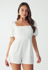 Witte romper met een vierkante halslijn en pofmouwen. Heeft verticale plooien in het bovenlijf en een getailleerde taille, afgewerkt met shorts.