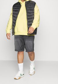 Felpa gialla a maniche corte sotto un gilet imbottito nero, abbinata a pantaloni corti grigi e sneakers bianche con dettagli verdi. Calze bianche visibili.