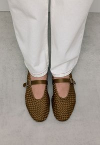 Ballerines tressées marron avec un bout arrondi, présentant une sangle latérale avec une boucle en argent, associées à un pantalon blanc retroussé.