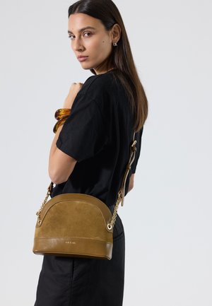 Femme aux longs cheveux foncés portant une robe noire et un bracelet en ambre, portant un sac à bandoulière marron en cuir et daim en forme de demi-lune.