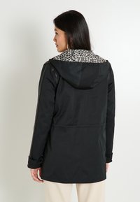 Mujer con cabello largo y oscuro, vestida con una chaqueta negra con capucha y forro de leopardo, de perfil contra un fondo claro y sencillo.