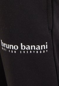 Juodas medvilnės audinys su lygiu tekstūra, kuriame iš šono spausdinta balta užrašai "bruno banani NOT FOR EVERYBODY".