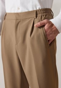 Pantalones beiges ajustados con frente plisado, cintura elástica y bolsillos laterales. Textura suave con un acabado de tela ligera.