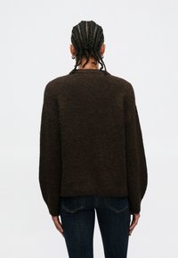 Bruine gebreide trui met een ontspannen pasvorm, verlaagde schouders en een textuurrijke afwerking. Gecombineerd met donkere jeans, gezien van achteren.