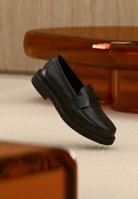 Μαύρο δερμάτινο μοκασίνι με χοντρή σόλα που ακουμπά σε ανοιχτόχρωμο χαλί, με ημιδιαφανή έπιπλα χρώματος κεχριμπαριού στο φόντο.