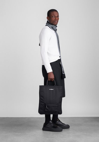 Mala tote preta em nylon com um bolso frontal com fecho e alças duplas; usada por uma pessoa com um suéter branco e calções pretos.