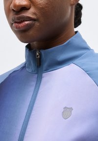 Posnetek od blizu osebe, ki nosi modro jopico z zadrgo in logotipom K-Swiss na prsih, prikazuje ustnice, brado in del vratu.