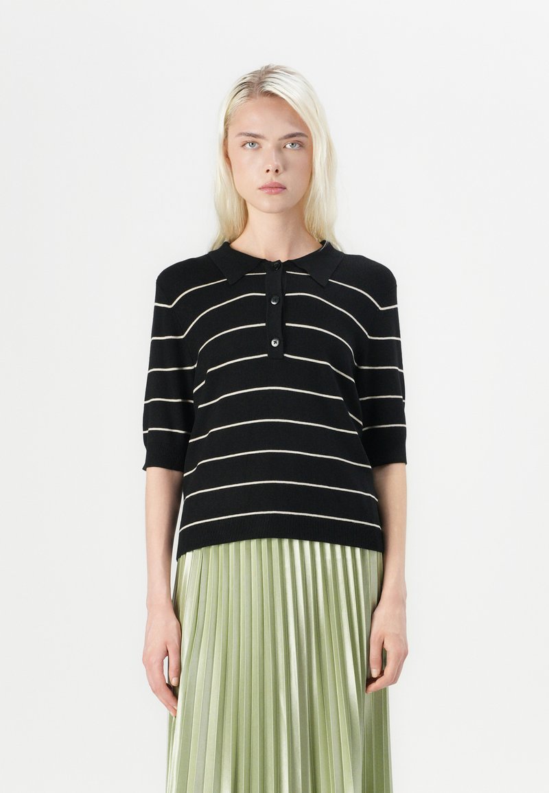 Maglione polo a righe bianche e nere in maglia, con maniche corte, abbinato a una gonna plissettata leggera verde chiaro e lucida, caratterizzata da una patta con bottoni.