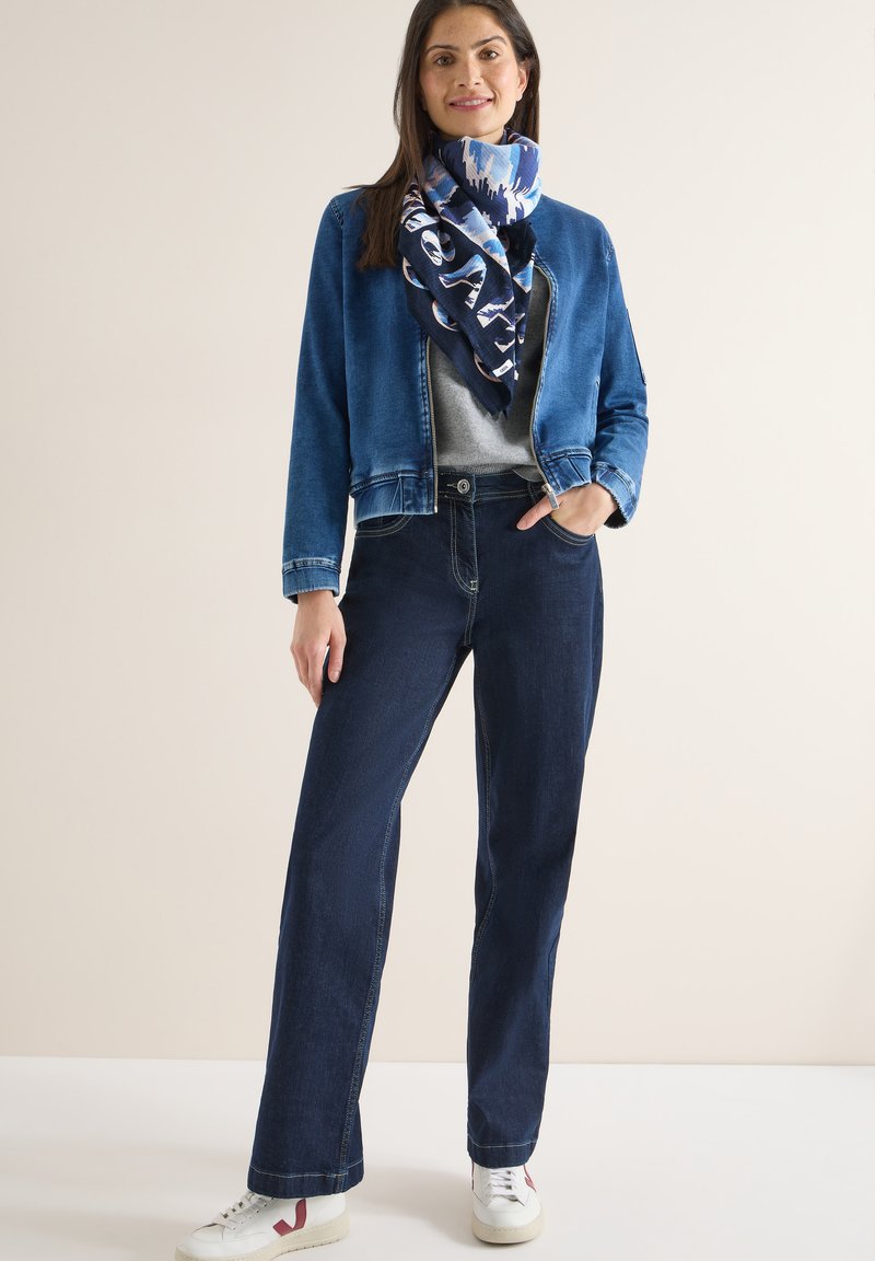 Femme debout portant une veste en denim bleue, une écharpe bleue à motifs, un haut gris, un jean bleu foncé et des baskets blanches devant un fond uni.