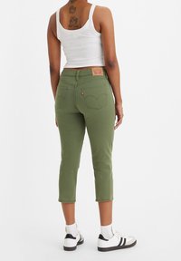 Oliwkowe przycięte dżinsy z denimu, pięć kieszeni, brązowa skórzana naszywka z tyłu. Modelka nosi białą bluzkę na ramiączkach i sneakersy.