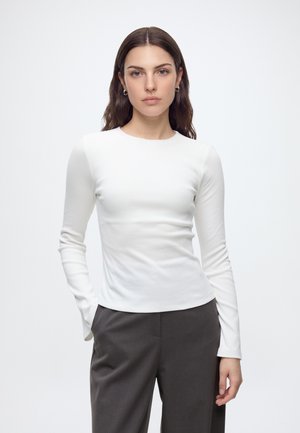 Femme aux longs cheveux bruns portant une chemise blanche à manches longues et un pantalon gris foncé, debout avec une main dans la poche devant un fond uni.