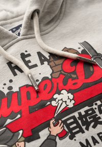 Felpa grigia con una grafica che presenta elementi rossi e neri, inclusi il testo "Superdry" e illustrazioni dettagliate. Accenti del cappuccio con cordini visibili.