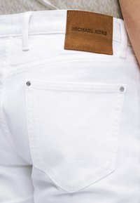 Λευκά τζιν από τζιν ύφασμα με καφέ δερμάτινη ετικέτα που φέρει την επιγραφή «MICHAEL KORS». Διαθέτουν πίσω τσέπες με ασημένια μεταλλικά στοιχεία και διπλή ραφή λεπτομέρειας.