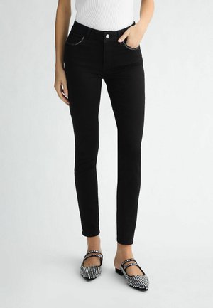 Person iført sorte slim fit jeans, en hvid ærmeløs top og sorte spidse flade sko med sølvnitter, stående på en lys baggrund.