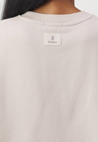 Vue de dos d'une personne portant un t-shirt beige clair à col rond avec une étiquette blanche Barbour cousue près du col.