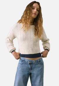 Maglione a maglia crema con texture a coste, maniche larghe e orlo blu navy. Abbinato a jeans alta vita blu chiaro con chiusura a bottone.