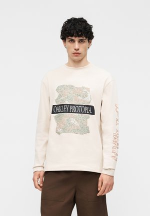 Mladý muž s kučeravými vlasmi, oblečený v krémovej dlhoročnej košeli s textom a grafikou "OAKLEY PROTOPIA", v kombinácii so hnedými nohavicami.