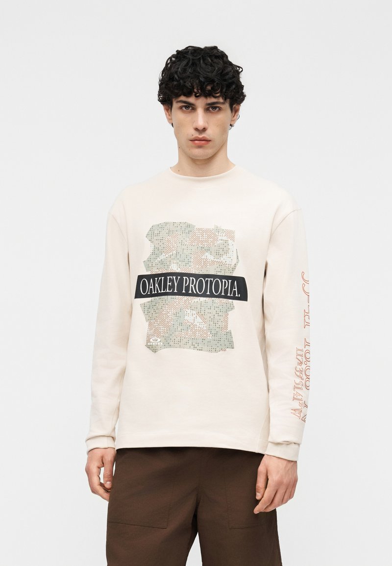 Νέος με κατσαρά μαλλιά, φοράει μια κρεμ μακρυμάνικη μπλούζα με το κείμενο και το γραφικό "OAKLEY PROTOPIA", συνδυασμένη με καφέ παντελόνια.