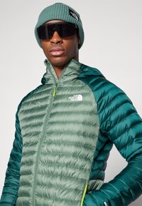 Gevoerde groene jas met een capuchon, uitgerust met een rits en een logo. gedragen met een geribde groene beanie en zonnebril. Gestructureerde stof en glans.