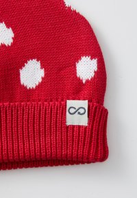 Rote gestrickte Mütze mit weißen Punkten, ausgestattet mit einem gerippten Saum und einem kleinen Stoffetikett mit einem Unendlichkeitssymbol. Die Textur wirkt weich.