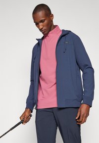 Veste zippée marine avec capuche, en tissu texturé et dotée de poches avant, portée sur un haut à manches courtes rose, tenue avec un club de golf noir.