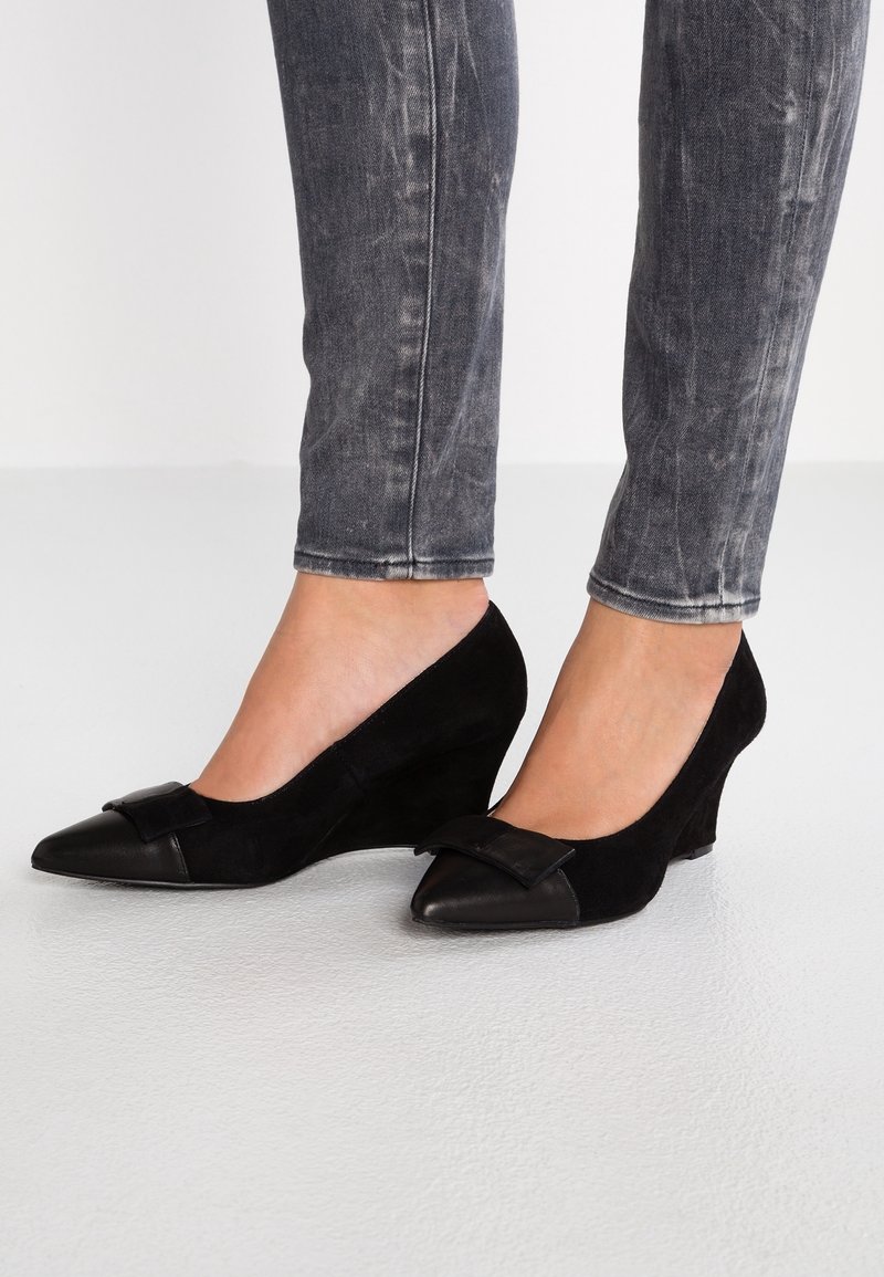 Pieds portant des compensées en daim noir à bouts pointus avec un détail nœud, associées à un jean skinny noir délavé.