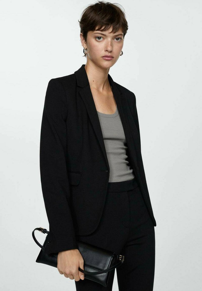 Black blazer over a gray fitted tank top, paired with black fitted pants. The model holds a small black clutch, featuring a strap.