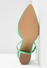 Chaussure à talon haut en daim vert à bout pointu, inclinée contre un mur blanc, montrant le détail de la semelle et du talon.
