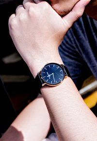Montre noire avec un boîtier rond rehaussé d'or, des index d'heures blancs et un bracelet noir, portée sur un poignet reposant sous un menton.