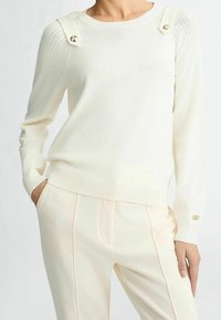 Bílý pletený svetr s žebrovanými rameny, ozdobnými knoflíky na bocích a úzkými manžetami. Kombinován s odpovídajícími světle zbarvenými kalhotami.