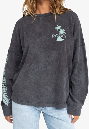 Woman wearing a loose dark gray long-sleeve shirt with palm tree logo and floral sleeve print, paired with light blue jeans.