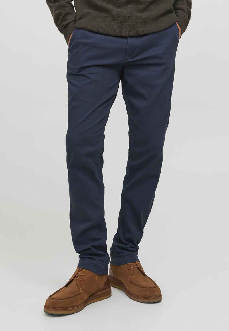Marineblaue Chinos aus weicher Baumwolle mit einem engen Schnitt. Kombiniert mit braunen Wildlederschuhen mit gesteppten Details und flacher Sohle.