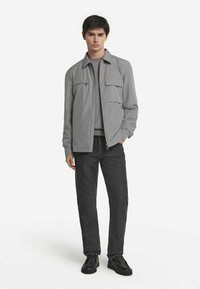 Ljusgrå zip-jacka med två framfickor och en texturerad grå skjorta under, kombinerad med mörka jeans och svarta skor.