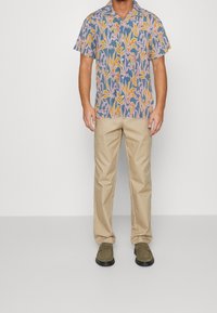 Kurzärmeliges, mehrfarbiges Hemd mit einem Blumenmuster in Blau, Gelb und Rosa, kombiniert mit beiger Hose und olivgrünen Schuhen.