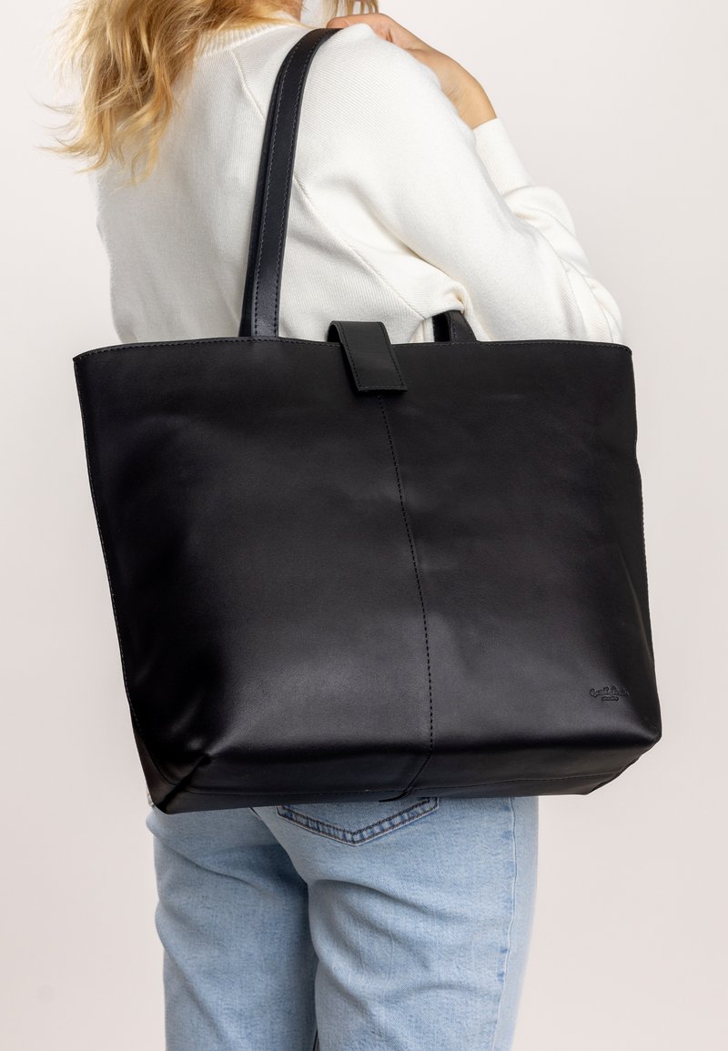 Zwarte leren tote bag met een gladde textuur, verticale naad en twee lange handvatten. Heeft een klein logo aan de zijkant en een centrale sluiting.