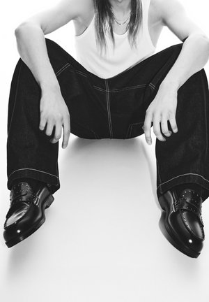 Černé leštěné kožené boty s nýty, nošené s volnými tmavými džínovými kalhotami a bílým tílkem, vsedě.