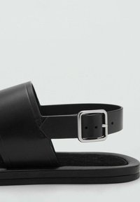 Sandalia de cuero negro que presenta una correa ancha, hebilla ajustable y una suela plana con una plantilla de textura suave. Diseño minimalista, sin patrones.
