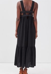 Robe longue noire avec larges bretelles, dos ouvert et ourlet à volants superposés. Le tissu semble léger avec une texture lisse.