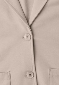 Veste en tissu beige avec deux boutons ronds et deux poches avant, avec un col et des revers cousus.