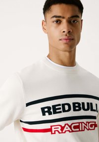 Suéter de punto blanco con el texto "RED BULL RACING" en negro y rojo. Diseño de cuello redondo con rayas contrastantes en el pecho. Textura suave.