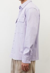 Camicia button-up di colore lilla chiaro, realizzata in cotone, con maniche lunghe, due tasche sul petto e orlo dritto. Indossata con pantaloni a motivi.