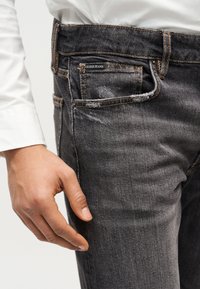 Δεξί χέρι χαλαρό δίπλα στην τσέπη σκουρόχρωμων ξεθωριασμένων τζιν Guess, που φοριέται από άτομο με λευκό μα κρυμάνικο πουκάμισο.