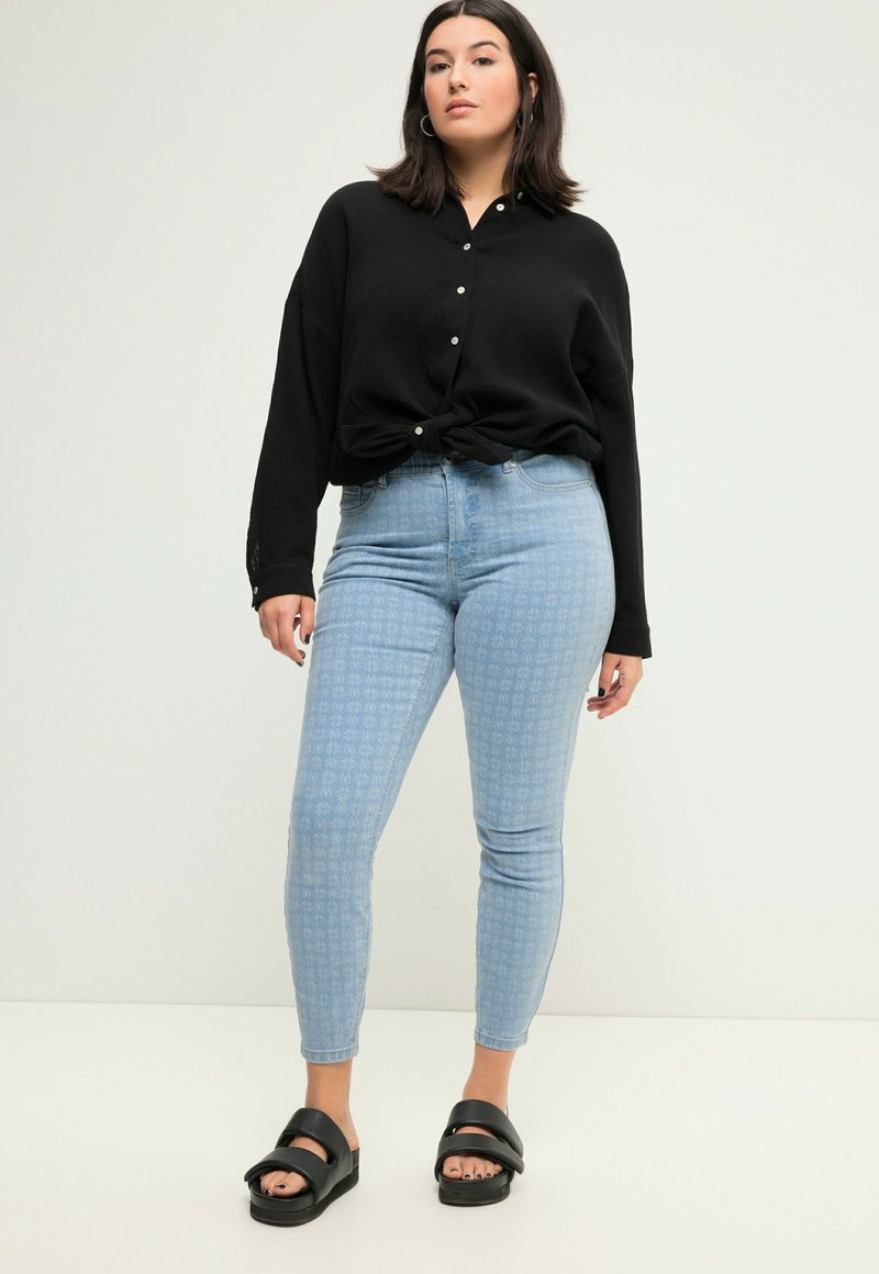 Camisa negra de botones atada en la cintura, jeans de talle alto azul claro con estampado y sandalias negras gruesas con dos tiras anchas.