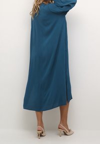 Langes, türkisfarbenes Kleid aus leichtem Stoff mit langen Ärmeln und einem Seitenschlitz. Kombiniert mit beigen High-Heels.