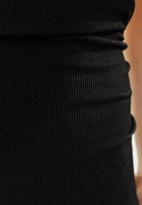 Svart ribbad tyg med vertikala linjer, mjuk textur, figurnära design. Har en rynkad midja som framhäver konturer och stretch.