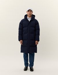 Uomo che indossa un lungo cappotto imbottito blu navy, un cappellino bianco, jeans e scarpe nere, in piedi contro uno sfondo chiaro e uniforme.