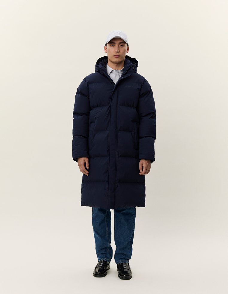 Uomo che indossa un lungo cappotto imbottito blu navy, un cappellino bianco, jeans e scarpe nere, in piedi contro uno sfondo chiaro e uniforme.