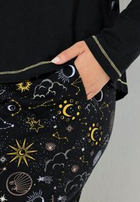 Černý top s dlouhým rukávem a zlatým prošíváním, spárovaný s černými kalhotami s nebeskými vzory v bílé, žluté a světle růžové na tmavém pozadí.